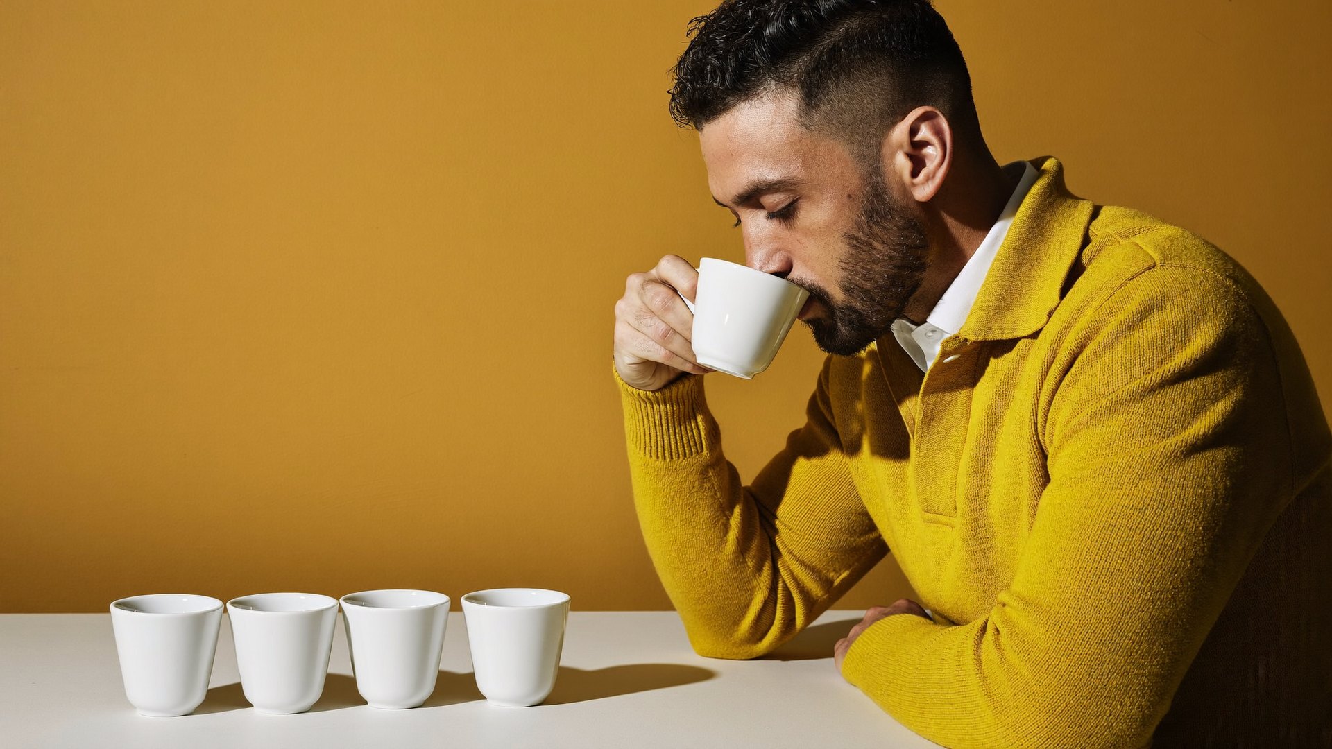 A man in a mustard jumper tasting from one of a row of small white cups