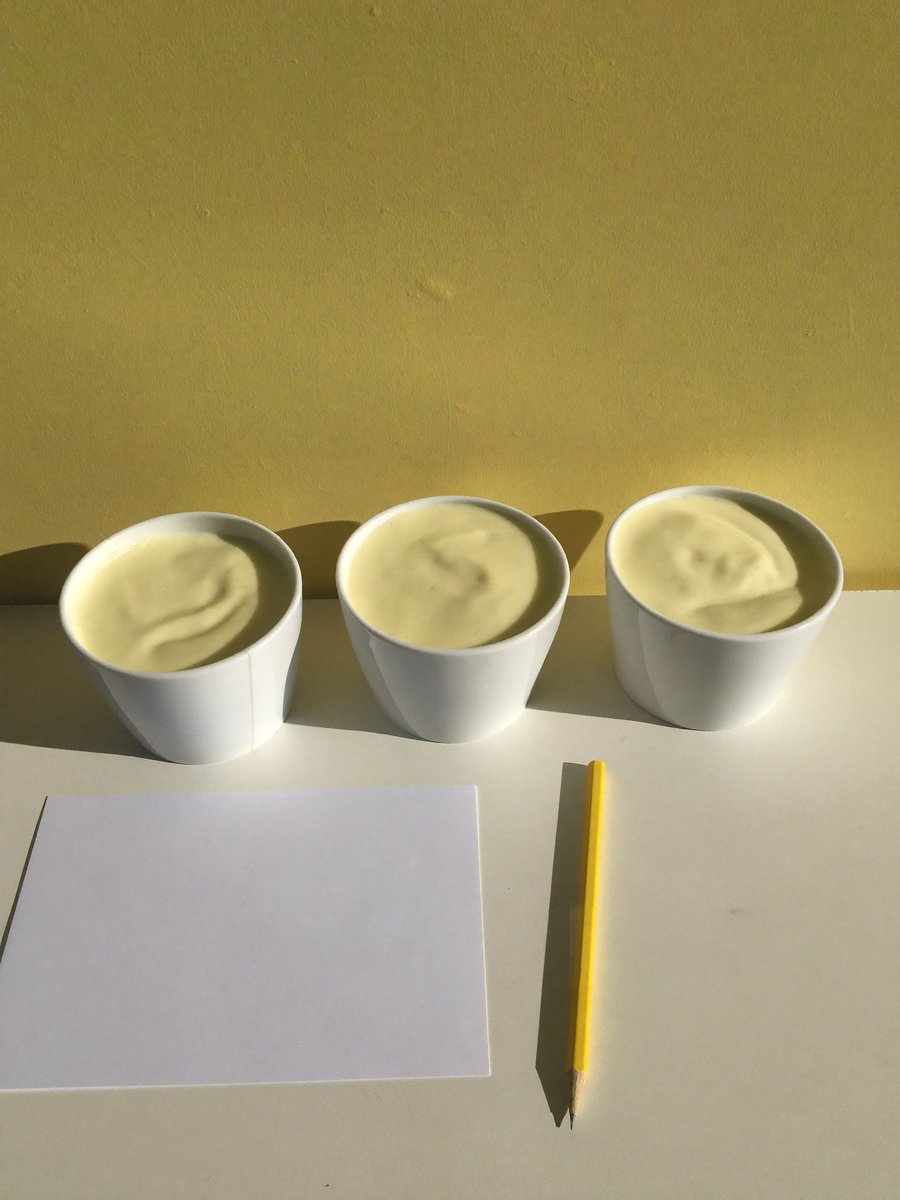 Three small white tasting cups of a pale smoothie beside a blank card and a pencil on a butter-yellow ground