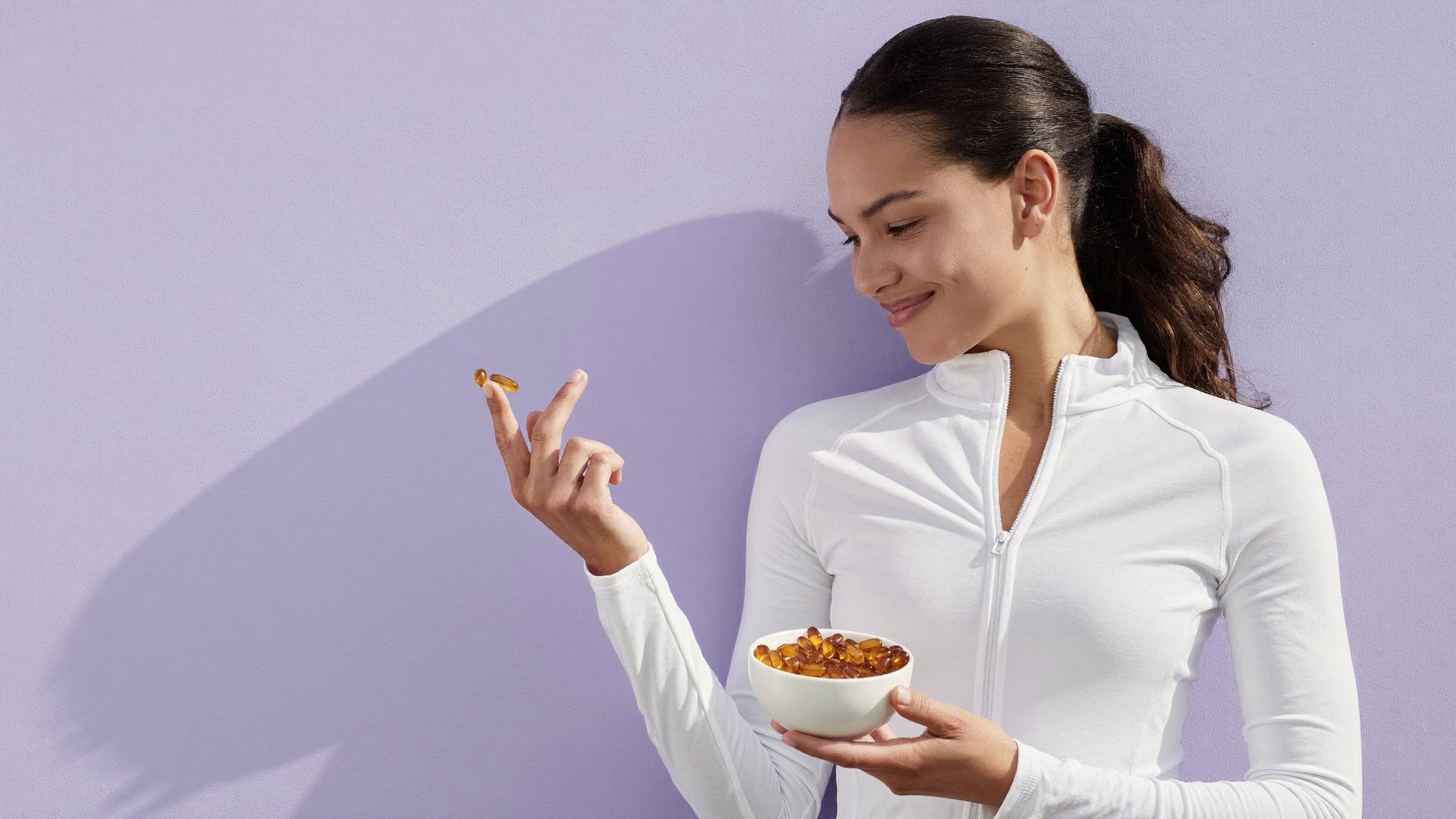 A woman in a white athletic top holding a small dish of softgels against a lilac wall
