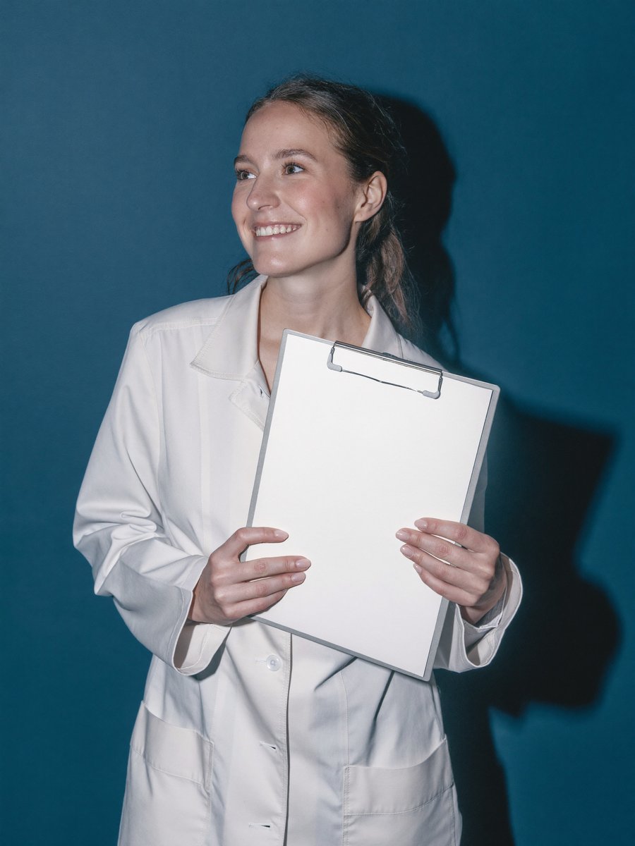 A clinical researcher in a white coat holding a clipboard against a deep teal wall