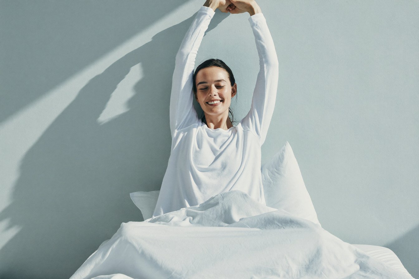 A woman sitting up in bed in the morning, stretching with a rested smile