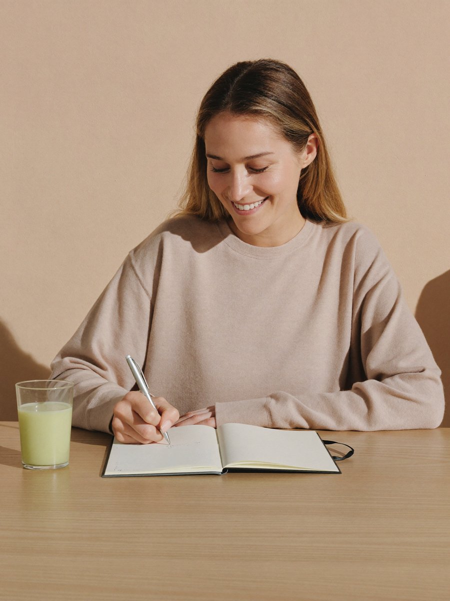 A woman writing in a notebook at her kitchen table beside a glass of pale green drink